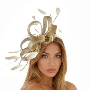 May include: A gold-colored headpiece with a bow and feather accents. The headpiece is made of a shimmering material and is worn on the head. The feathers are a light cream color. The background is white.