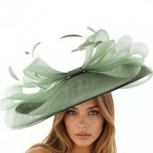 May include: A sage green hat with a wide brim and a large bow. The hat is adorned with delicate feathers and sheer fabric, creating a layered effect. The hat is a stylish accessory.
