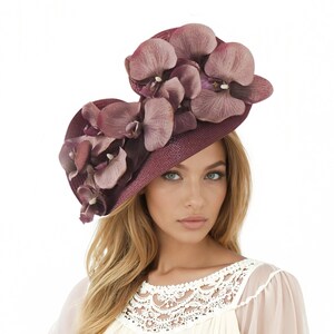 May include: A burgundy hat with a wide brim, decorated with large, mauve-coloured orchid blossoms. The hat has a textured appearance and is designed for special occasions. The hat is a statement piece, perfect for weddings or formal events.
