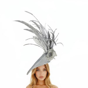 May include: A silver-colored hat with a dramatic, asymmetrical design. The hat features a wide brim and is adorned with large, feathery plumes and a decorative flower. The hat is designed for special occasions.