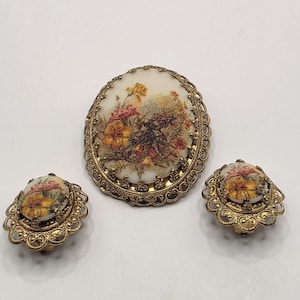 May include: A vintage jewelry set featuring a large oval brooch and matching clip-on earrings. The set has a gold-tone filigree frame surrounding a floral design in shades of yellow, red, and pink on a cream background.