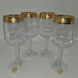 May include: Four clear wine glasses with gold rims and decorative etching. Each glass has a long stem and a round base. The gold rim features an ornate design. The glasses are labeled "CELLINI" on the base.