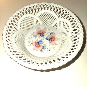 May include: A white porcelain basket with a delicate openwork design. The basket holds a small, round dish decorated with pink and blue floral patterns. The overall aesthetic is vintage and elegant, suitable for display or serving.