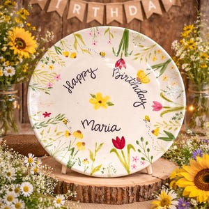 May include: A round white ceramic plate featuring hand-painted floral motifs including yellow sunflowers, red tulips, and small bees. The text "Happy Birthday" and the name "Maria" are written in black script in the center of the plate.