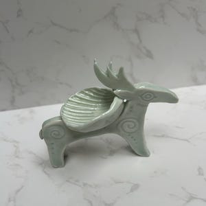 May include: A pale green ceramic figurine shaped like a stylised deer with a shell-like dish resting on its back. The deer features etched spiral patterns on its body and legs, with a smooth, glossy finish and a pointed head with antlers.