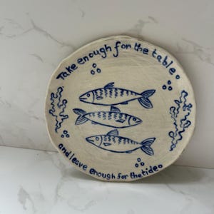 May include: A round, off-white ceramic plate featuring hand-drawn blue illustrations of three fish, seaweed, and bubbles. The text "Take enough for the table and leave enough for the tide" is written in blue along the curved edge of the plate.