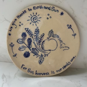 May include: A round ceramic plate with a cream-coloured glaze and blue hand-drawn illustrations of wheat, berries, an apple, and a sun. The text reads: We give thanks to Earth and Sun. For this harvest is our plenty won.