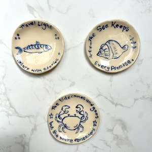 May include: Three small, round, off-white ceramic trinket dishes with hand-painted blue illustrations and text. One features a fish with 'Travel Light, return with stories', another a seashell with 'The Sea Keeps Every Promise', and one a crab with 'The tide comes and goes but the shore remembers'.