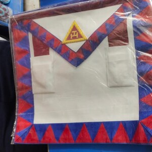 Könnte beinhalten: Weißer Freimaurer-Schurz mit rotem und blauem Besatz. Der Schurz zeigt ein goldenes Dreieck mit den Buchstaben 'F.H.' darin. Der Schurz hat zwei Taschen.