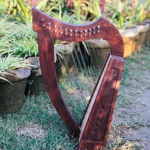 May include: A wooden Celtic harp with a carved design on the soundboard. The harp is brown and has a natural finish. It is sitting on a bed of green grass.