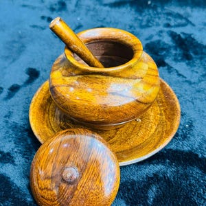 Könnte beinhalten: Eine Holzschale mit Deckel und einem Holzlöffel. Die Schale ist braun und hat eine natürliche Holzoptik. Der Deckel ist rund und hat einen Knopf oben. Der Löffel ist lang und dünn.