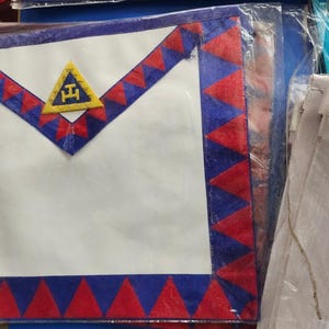 Puede incluir: Un delantal blanco masónico con un borde azul y rojo. El delantal tiene un triángulo amarillo con las letras 'F.H.' en el centro.