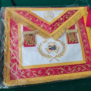 Könnte beinhalten: Weißer Freimaurer-Schurz mit rotem und goldenem Besatz. Der Schurz zeigt ein golden gesticktes Design mit Zirkel und Winkel, einem Kranz und einem Dreieck mit einem Auge. Der Schurz hat zwei goldene Quasten.