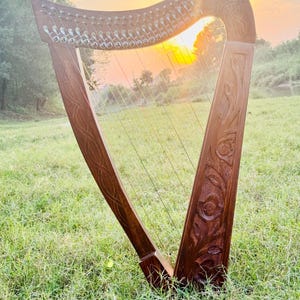 以下が含まれることがあります： 複雑な彫刻と弦を備えた木製のハープ。夕焼けを背景に、草の生えた屋外に設置されています。ハープは、職人技を示す詳細な花柄とケルトノットのデザインが特徴です。楽器は直立しており、演奏する準備ができています。
