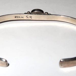 May include: A silver cuff bracelet with an open design. The interior surface is stamped with the text STERLING and the letters SS. The bracelet features a smooth, polished metallic finish and a rounded band that tapers toward the open ends.