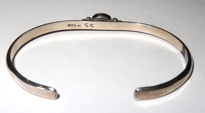 May include: A silver cuff bracelet with an open design. The interior surface is stamped with the text STERLING and the letters SS. The bracelet features a smooth, polished metallic finish and a rounded band that tapers toward the open ends.