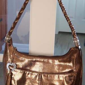 Puede incluir: Un bolso de mano de cuero de color dorado met&aacute;lico con una correa de cuero trenzada marr&oacute;n y un dije de coraz&oacute;n plateado.
