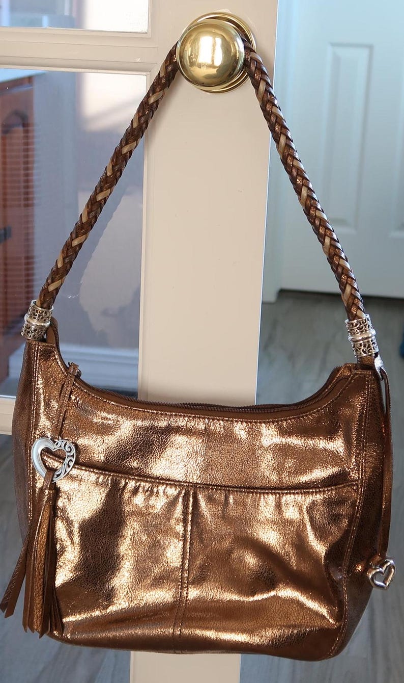 Puede incluir: Un bolso de mano de cuero de color dorado met&aacute;lico con una correa de cuero trenzada marr&oacute;n y un dije de coraz&oacute;n plateado.