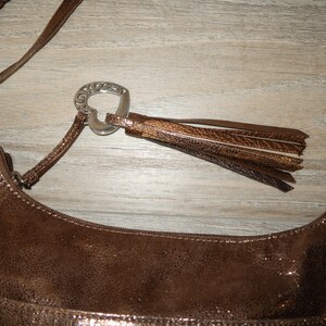 Puede incluir: Un bolso de mano de cuero marr&oacute;n con un cierre en forma de coraz&oacute;n plateado y una correa trenzada con detalles plateados. El bolso tiene un borla.