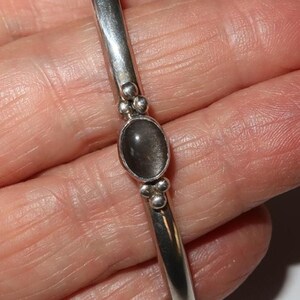 May include: A silver bangle bracelet featuring a smooth, polished band and a central oval-shaped dark gray stone setting. The stone is flanked by small decorative silver spheres on both the top and bottom, creating a balanced and detailed design.