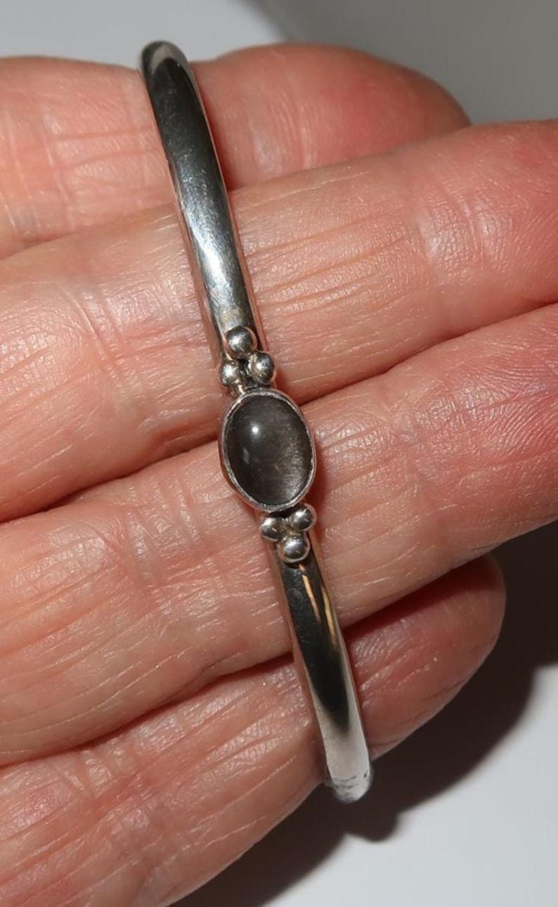 May include: A silver bangle bracelet featuring a smooth, polished band and a central oval-shaped dark gray stone setting. The stone is flanked by small decorative silver spheres on both the top and bottom, creating a balanced and detailed design.