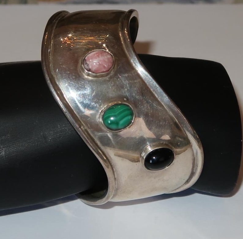 May include: A wide silver cuff bracelet featuring three oval-shaped cabochon gemstones set in a row. The stones include a pink mottled stone, a green banded malachite, and a smooth black stone, all encased in a polished, curved silver band.