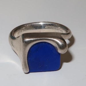 May include: A silver ring with a rectangular, deep blue stone. The ring has a unique, curved design with the words "TIFFANY & CO. 925" and "BENNETT SPAIN" engraved on the band.