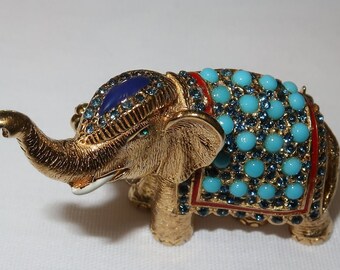May include: A gold-colored elephant figurine with blue and turquoise rhinestones. The elephant is wearing a decorative blue and turquoise patterned cloth.