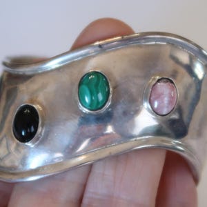 May include: A wide, polished silver cuff bracelet featuring a wavy, undulating edge. The bracelet is set with three smooth, oval-shaped cabochon gemstones arranged horizontally: a solid black stone, a green malachite stone, and a pale pink stone.