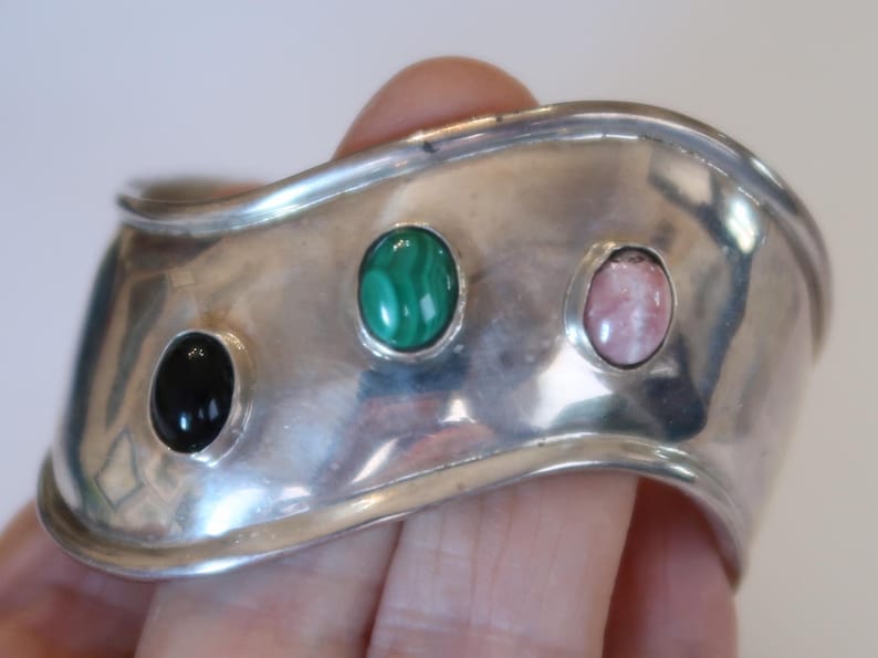 May include: A wide, polished silver cuff bracelet featuring a wavy, undulating edge. The bracelet is set with three smooth, oval-shaped cabochon gemstones arranged horizontally: a solid black stone, a green malachite stone, and a pale pink stone.