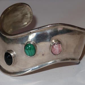 May include: A wide, polished silver cuff bracelet featuring a wavy, undulating edge. The bracelet is set with three smooth, oval-shaped cabochon stones: one solid black stone, one green and black banded malachite, and one pale pink stone.