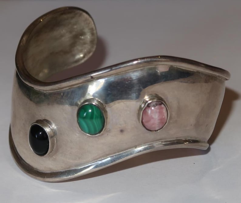 May include: A wide, polished silver cuff bracelet featuring a wavy, undulating edge. The bracelet is set with three smooth, oval-shaped cabochon stones: one solid black stone, one green and black banded malachite, and one pale pink stone.
