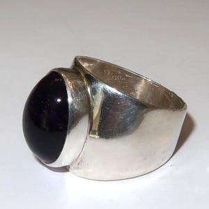 May include: A wide band ring crafted from polished silver metal, featuring a large, smooth, oval-shaped dark purple cabochon gemstone set securely in a bezel mounting. The ring has a solid, substantial construction with a reflective surface.