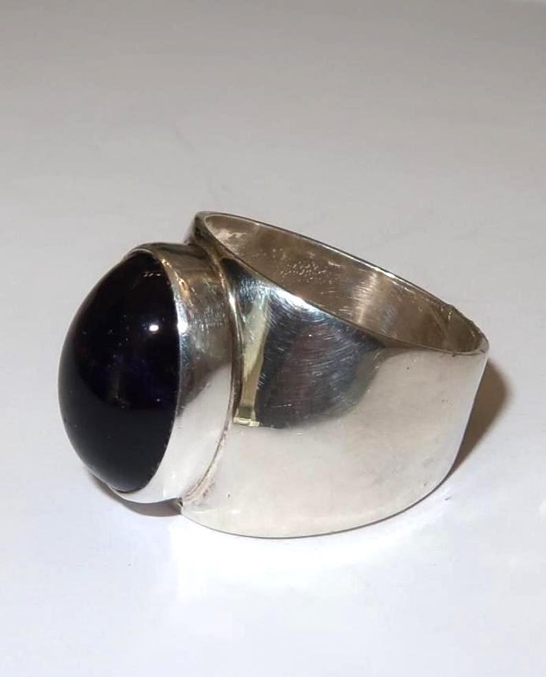 May include: A wide band ring crafted from polished silver metal, featuring a large, smooth, oval-shaped dark purple cabochon gemstone set securely in a bezel mounting. The ring has a solid, substantial construction with a reflective surface.