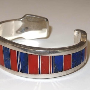 May include: Silver cuff bracelet with alternating red and blue inlays. The bracelet features a rectangular design with vertical stripes of red and blue, separated by thin white lines. The metal has a polished finish.