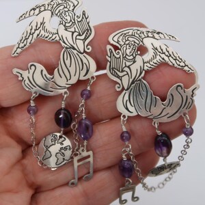 May include: A pair of silver earrings featuring angel figures playing harps. The earrings have dangling amethyst beads, musical notes, and a small globe charm. The angel wings are detailed with etched lines.