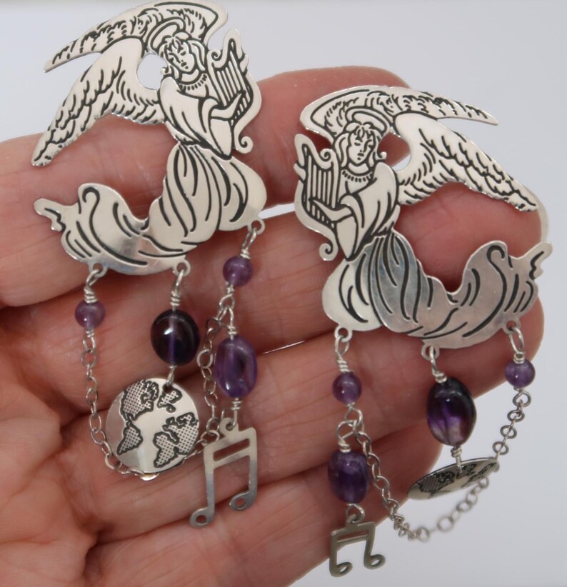May include: A pair of silver earrings featuring angel figures playing harps. The earrings have dangling amethyst beads, musical notes, and a small globe charm. The angel wings are detailed with etched lines.