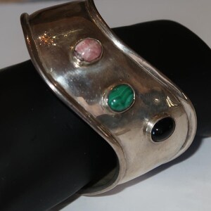 May include: A wide silver cuff bracelet featuring a smooth, polished metallic surface. The bracelet is set with three distinct oval-shaped cabochon gemstones: a pink stone, a green banded malachite stone, and a solid black stone.