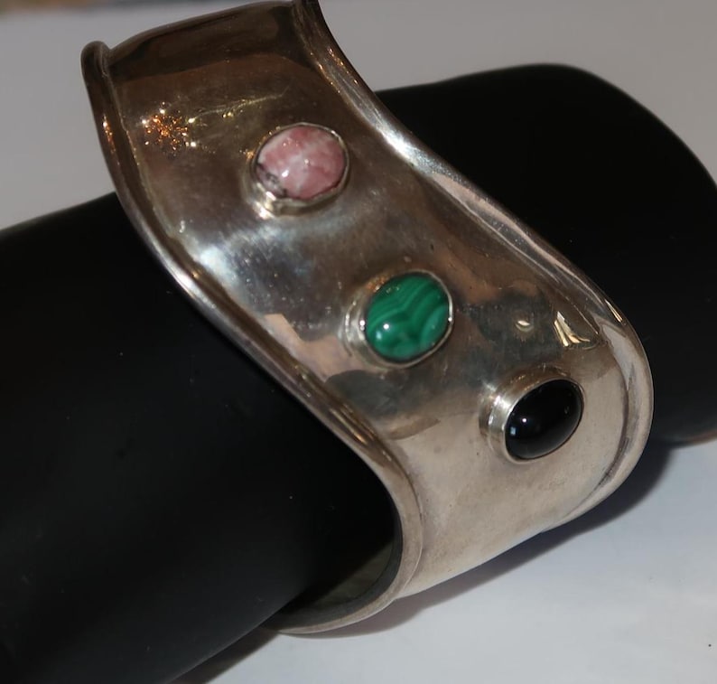 May include: A wide silver cuff bracelet featuring a smooth, polished metallic surface. The bracelet is set with three distinct oval-shaped cabochon gemstones: a pink stone, a green banded malachite stone, and a solid black stone.