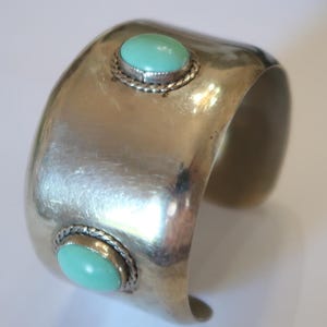 May include: A wide, polished silver-tone cuff bracelet featuring two smooth, round, light green cabochon stones. Each stone is set within a decorative twisted rope border, creating a distinct contrast against the smooth, curved metal surface.