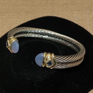 May include: Silver-tone cuff bracelet with a twisted rope design. The bracelet features gold accents and two oval, light blue gemstones. Small, square, dark blue gemstones are also present.