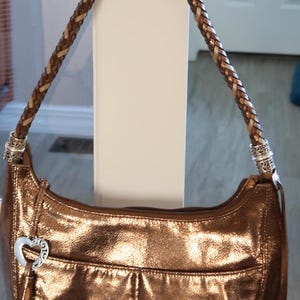 Puede incluir: Un bolso de mano de color dorado met&aacute;lico con una correa de cuero trenzada y un dije en forma de coraz&oacute;n. El bolso tiene un cierre de solapa y un peque&ntilde;o bolsillo en la parte delantera.