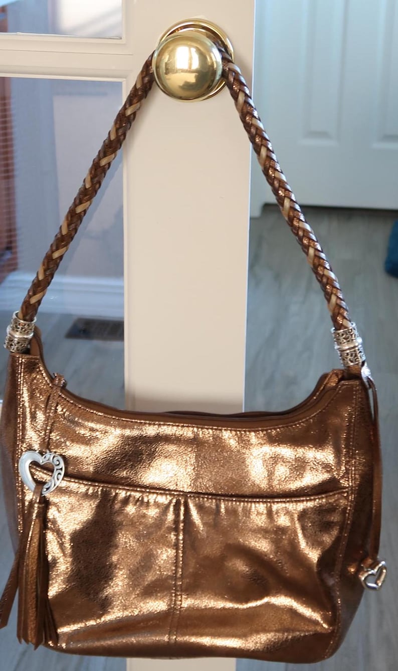 Puede incluir: Un bolso de mano de color dorado met&aacute;lico con una correa de cuero trenzada y un dije en forma de coraz&oacute;n. El bolso tiene un cierre de solapa y un peque&ntilde;o bolsillo en la parte delantera.