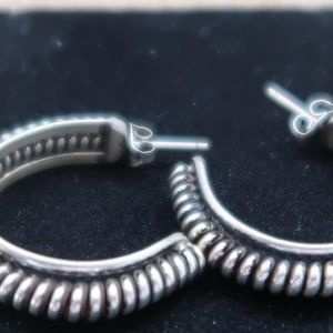 May include: A pair of silver hoop earrings. The earrings have a textured, ridged design on the outer edge and a smaller, detailed inner band. The earrings are displayed on a dark surface.