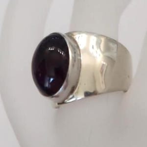 May include: A silver ring featuring a large, smooth, oval-shaped dark purple gemstone set in a polished metal bezel. The band of the ring is wide, crafted from a reflective silver-toned metal with a slightly hammered or uneven surface texture.