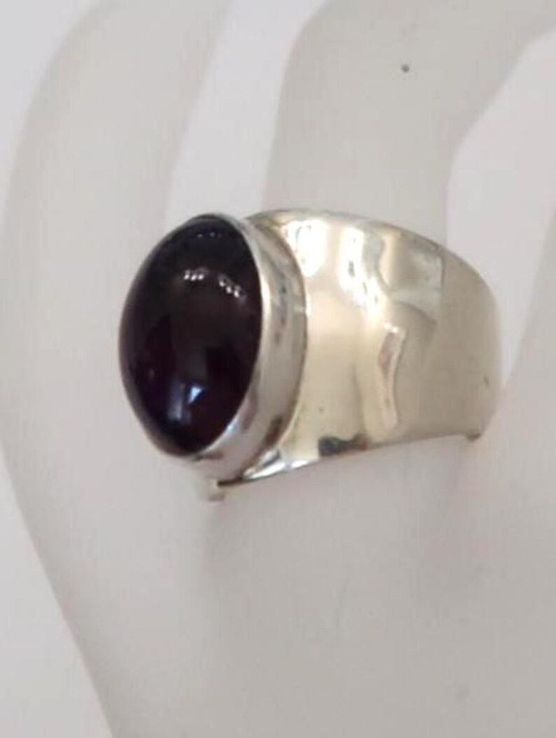 May include: A silver ring featuring a large, smooth, oval-shaped dark purple gemstone set in a polished metal bezel. The band of the ring is wide, crafted from a reflective silver-toned metal with a slightly hammered or uneven surface texture.