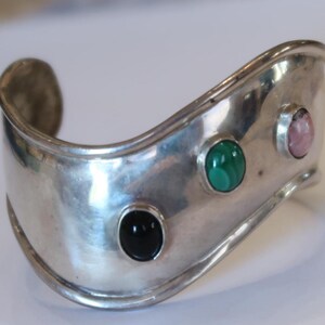 May include: A wide, polished silver cuff bracelet featuring a wavy, undulating edge. The metal surface is set with three smooth, oval-shaped cabochon gemstones arranged in a row: a solid black stone, a green malachite stone, and a pale pink stone.