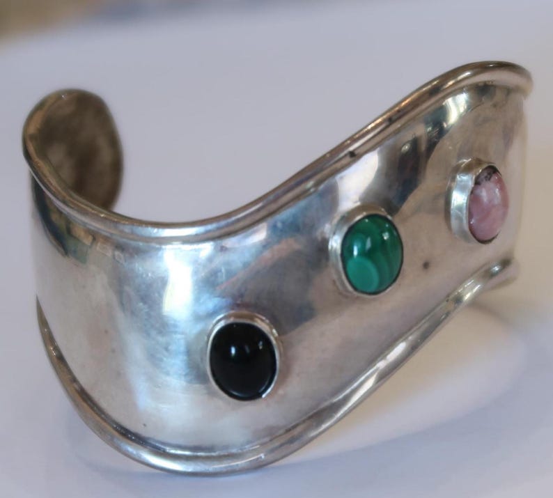 May include: A wide, polished silver cuff bracelet featuring a wavy, undulating edge. The metal surface is set with three smooth, oval-shaped cabochon gemstones arranged in a row: a solid black stone, a green malachite stone, and a pale pink stone.