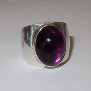 May include: A wide silver band ring featuring a large, smooth, oval-shaped deep purple cabochon gemstone set in the center. The metal band has a polished, reflective finish and a substantial, thick profile that tapers slightly toward the back.