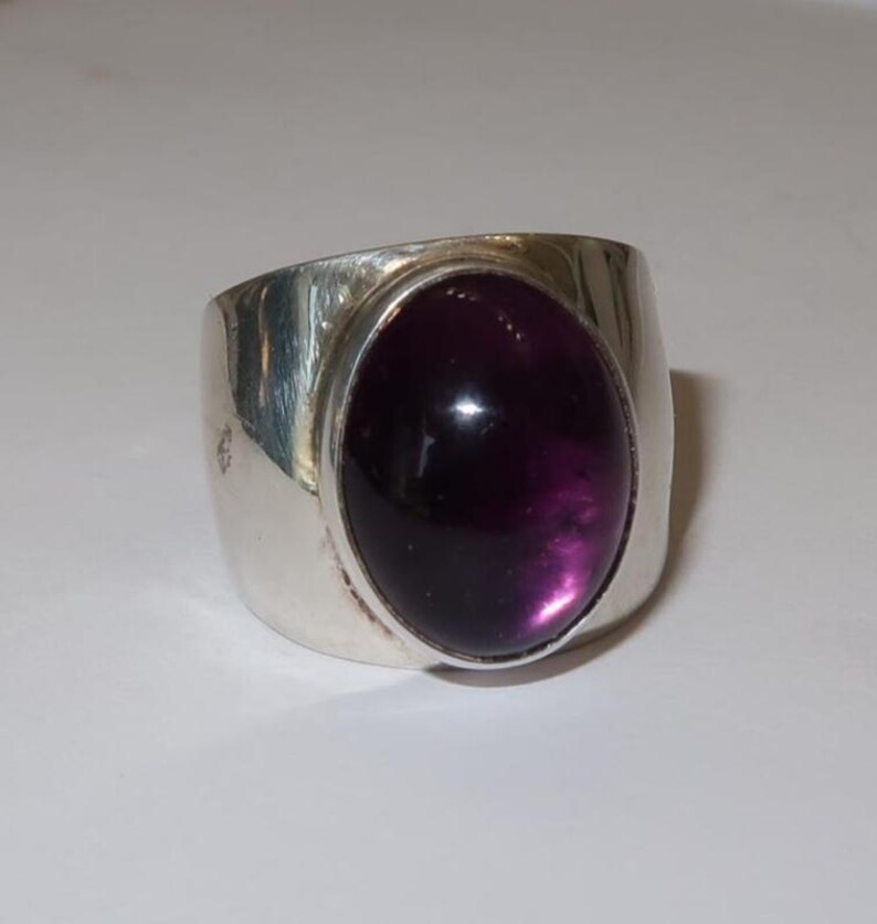 May include: A wide silver band ring featuring a large, smooth, oval-shaped deep purple cabochon gemstone set in the center. The metal band has a polished, reflective finish and a substantial, thick profile that tapers slightly toward the back.
